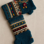 A pair of handmade crochet fingerless gloves in teal blue, decorated with colorful geometric patterns and embroidery in beige, brown, red, yellow, and blue, displayed on a light beige background.