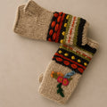 Hand-knitted winter mittens in a warm beige wool, featuring colorful striped and patterned cuffs with red, orange, green, yellow, and black yarn, and embroidered flowers on the front.